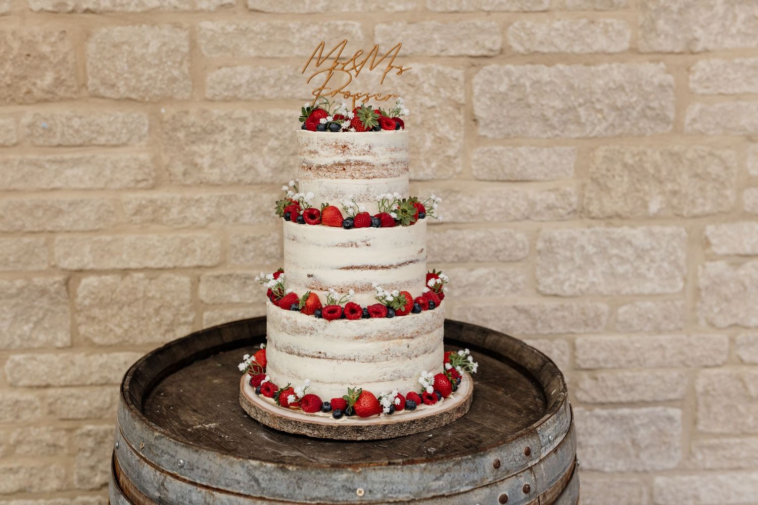 A tiered white wedding cake decorated with fresh strawberries and cream being assembled on a stone pedestal.