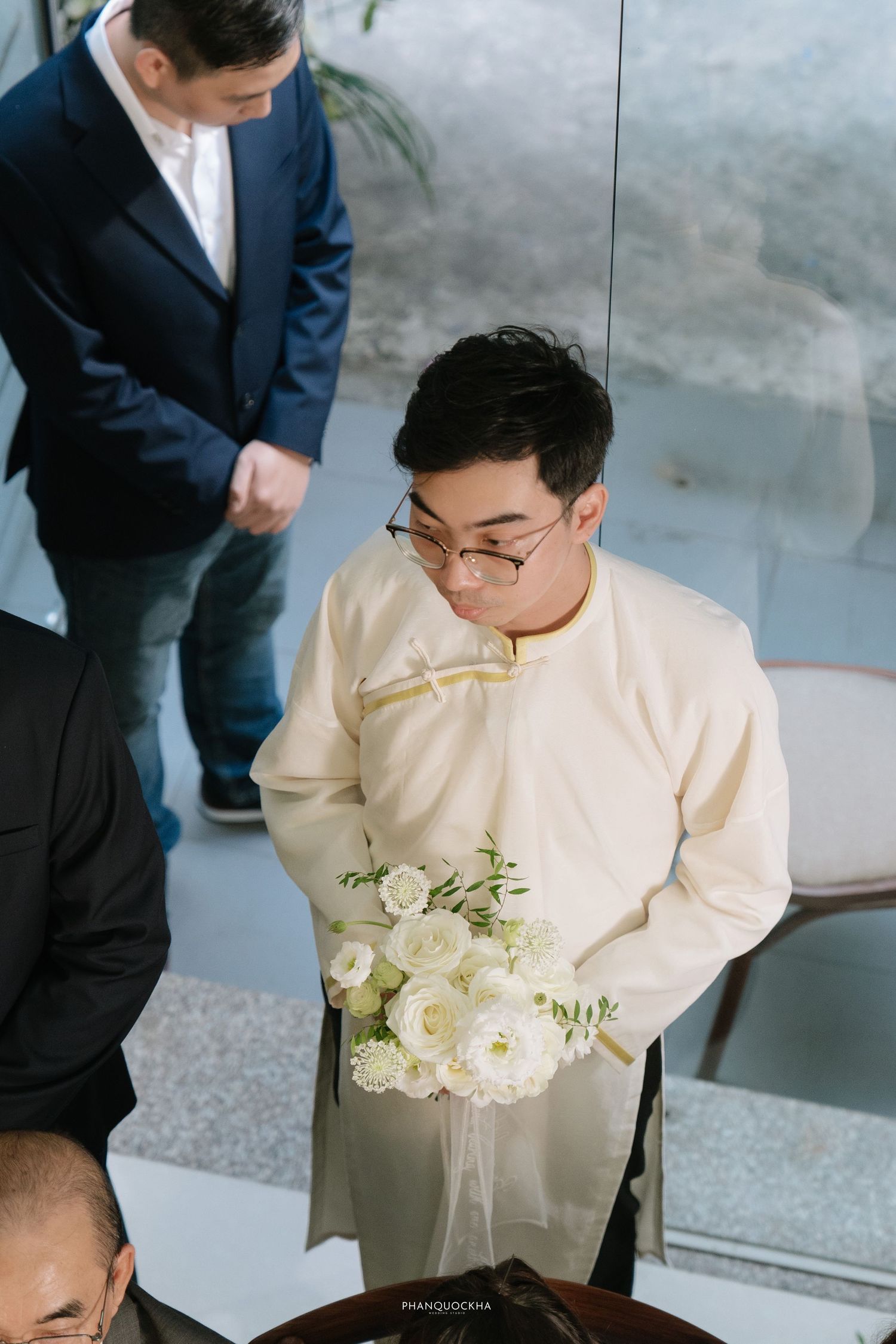 Groom in cream ao dai holding white flower bouquet viewed from above at a traditional Vietnamese wedding.