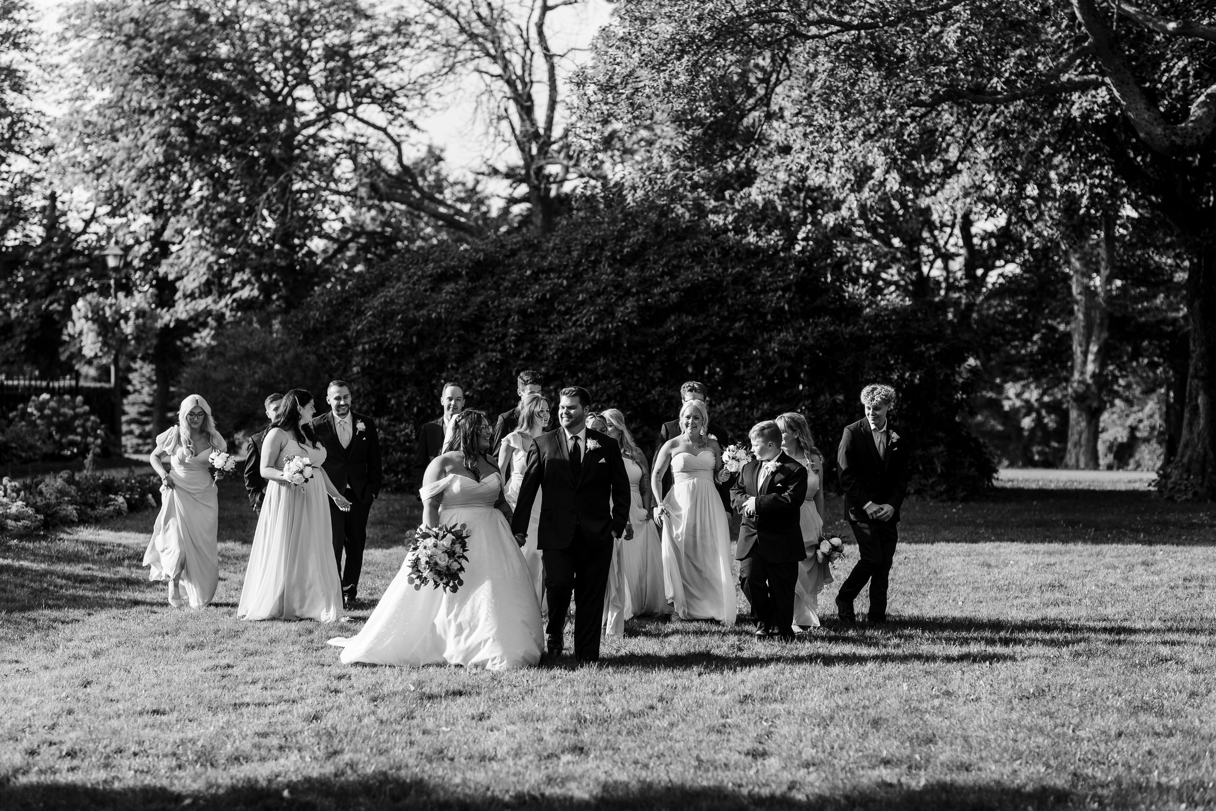 Wedding party in formal attire walking through a scenic garden path in black and white photography.