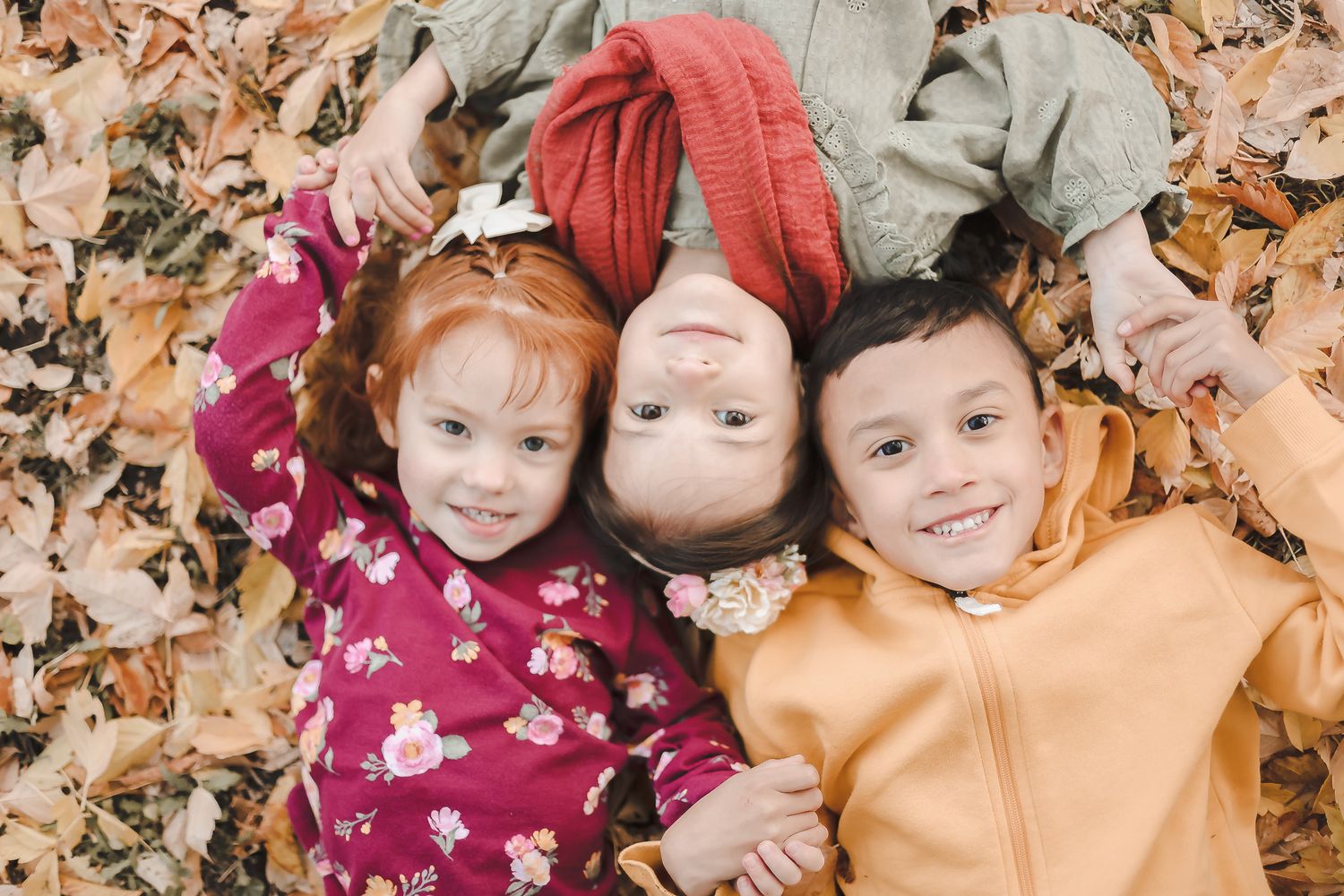 Three cheerful children lying in colorful autumn leaves while wearing fall sweaters.