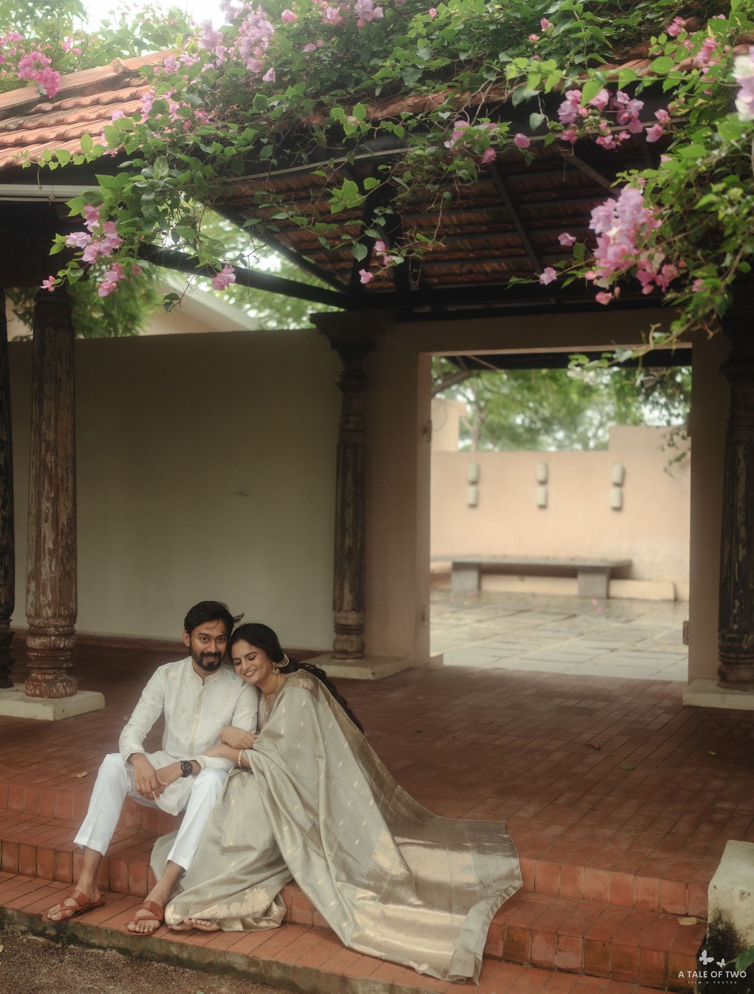 A couple sitting together under a romantic pergola covered in pink flowering vines at a peaceful outdoor setting.
