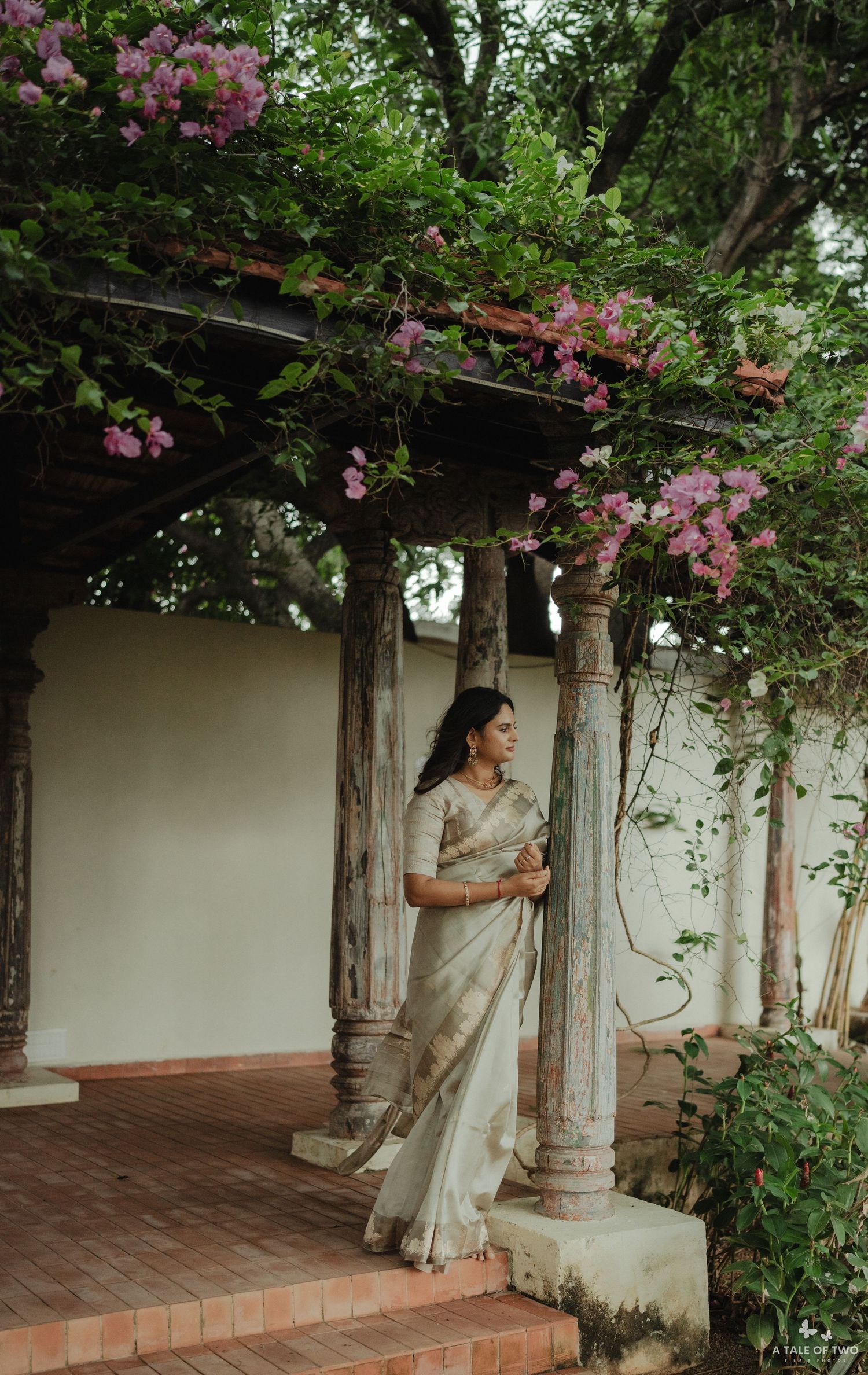 A person in an elegant cream sari stands under a covered porch with pink flowering vines and ornate columns.