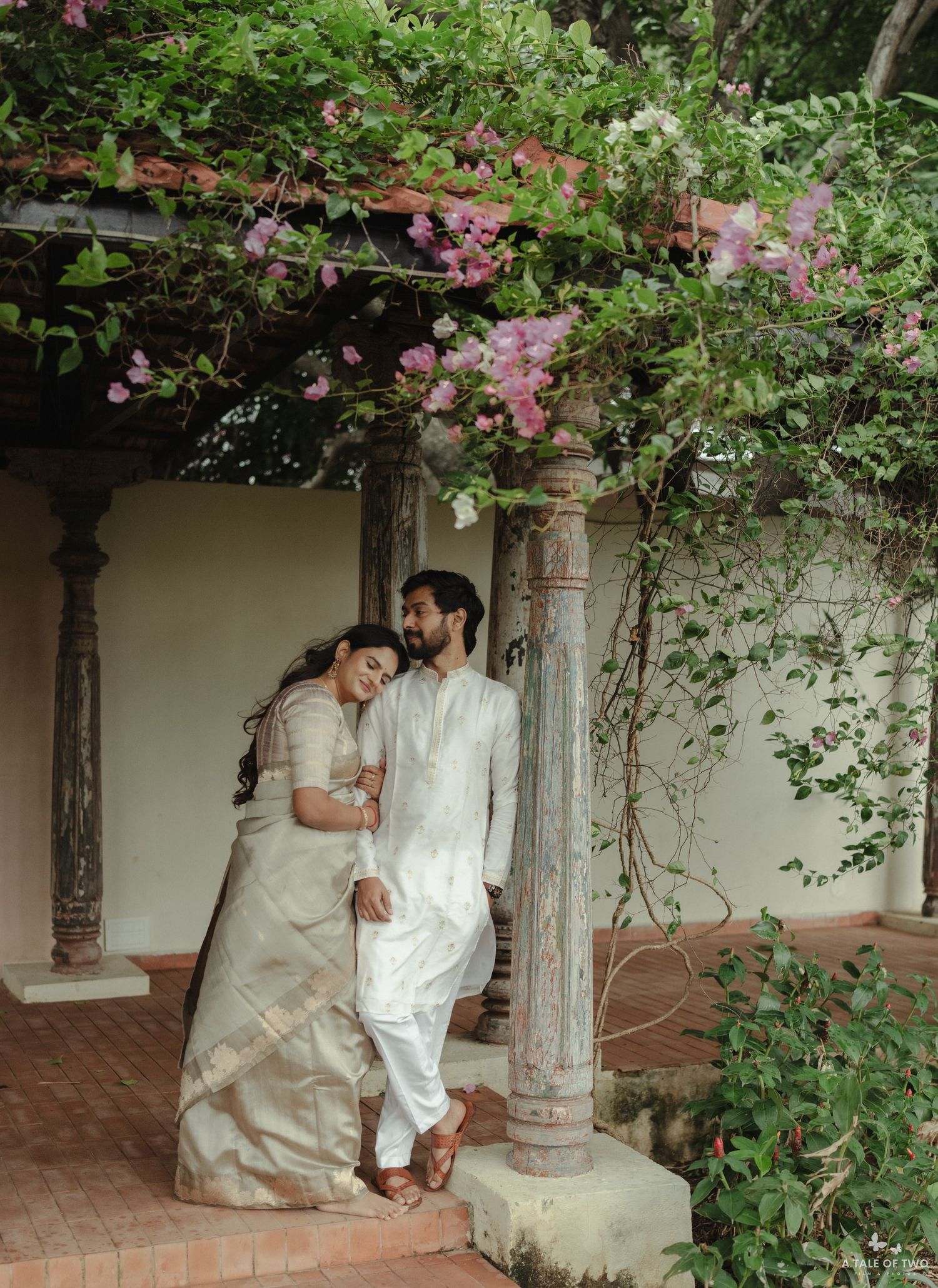Couple in elegant white attire embraces in a romantic garden walkway with pink flowers and stone pillars.