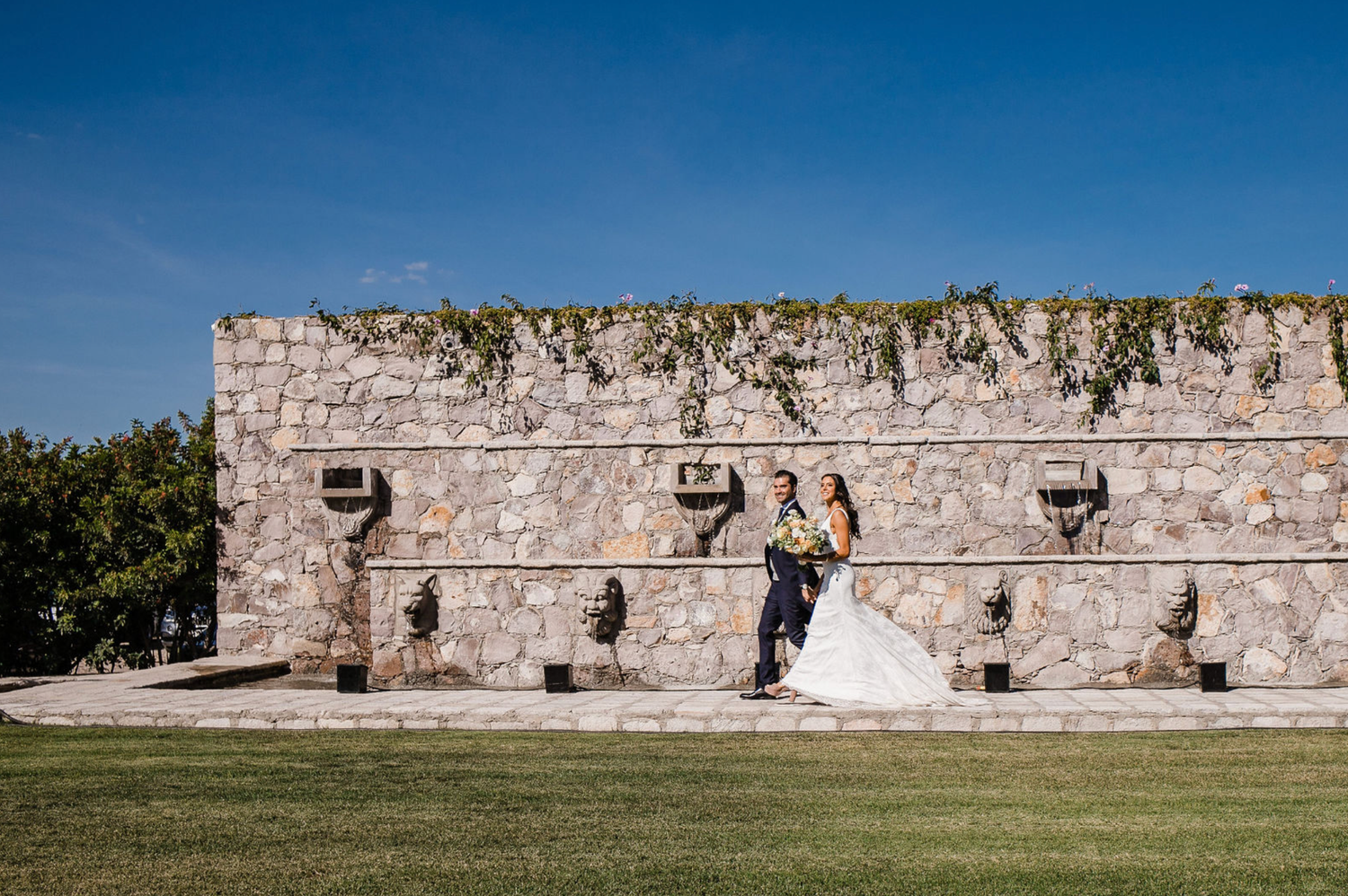 Wedding couple poses along a historic stone wall with expansive green lawn.