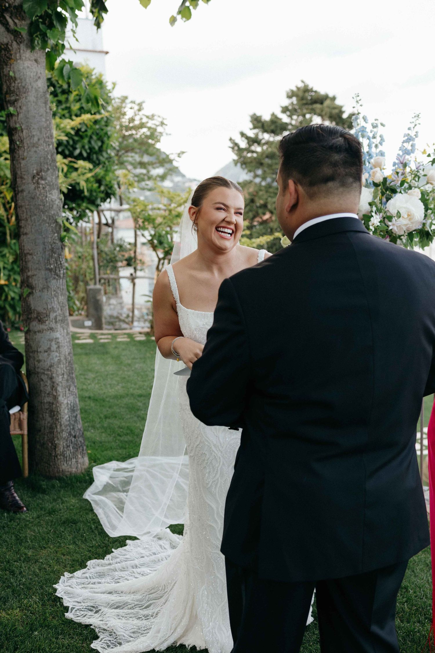 A joyful outdoor wedding scene with a bride in a lace gown and groom in a black suit standing on a lawn surrounded by trees.