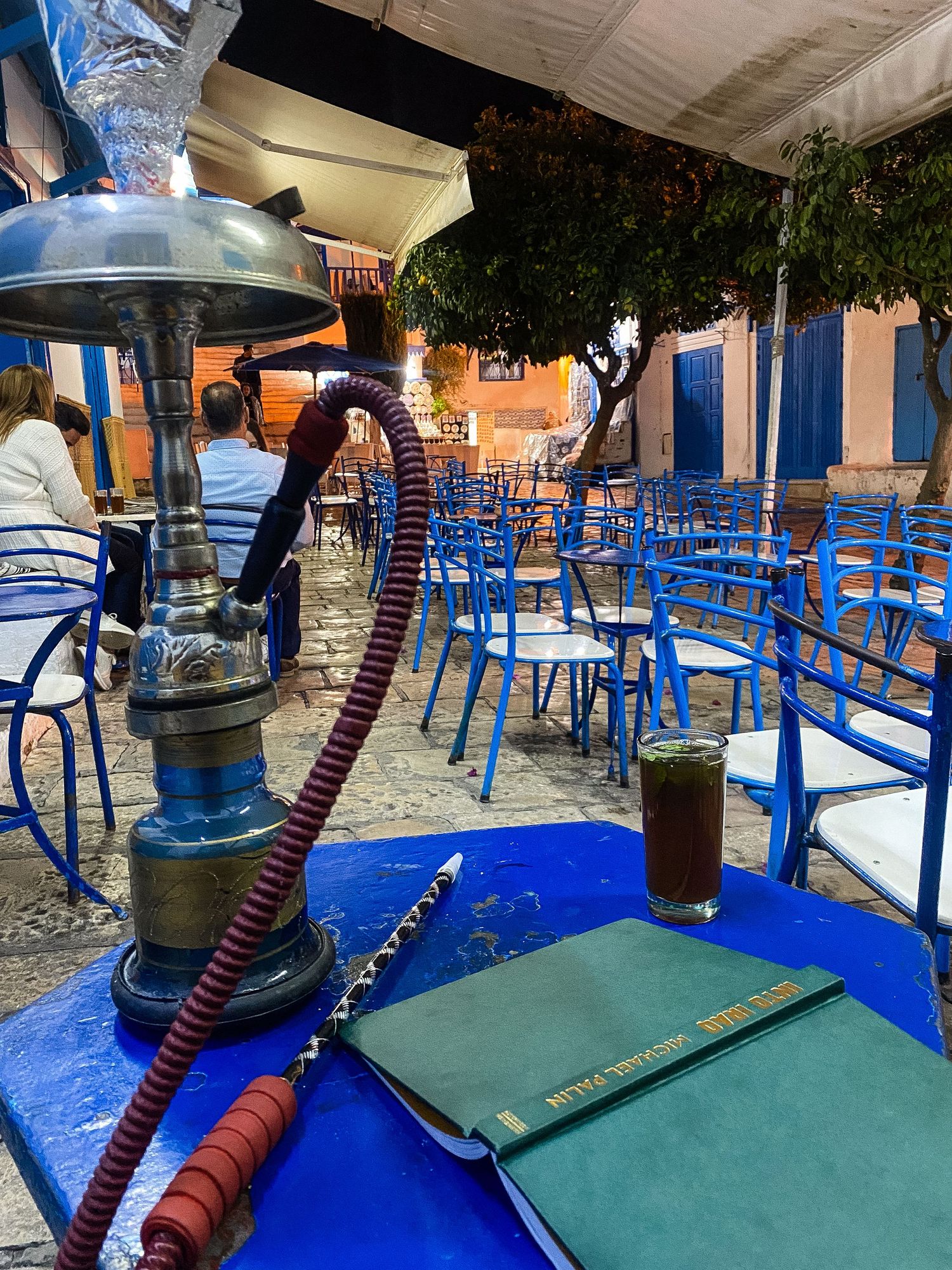 Hookah and drinks sit on bright blue tables at an outdoor Mediterranean cafe with chairs arranged for evening service.