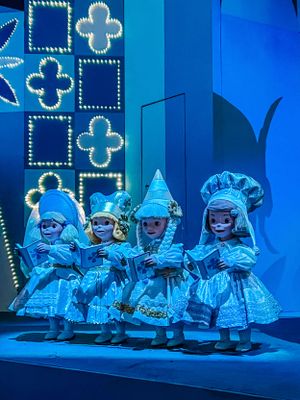 Performers in bright blue illuminated costumes and headpieces stand on stage in front of glowing geometric backdrop.