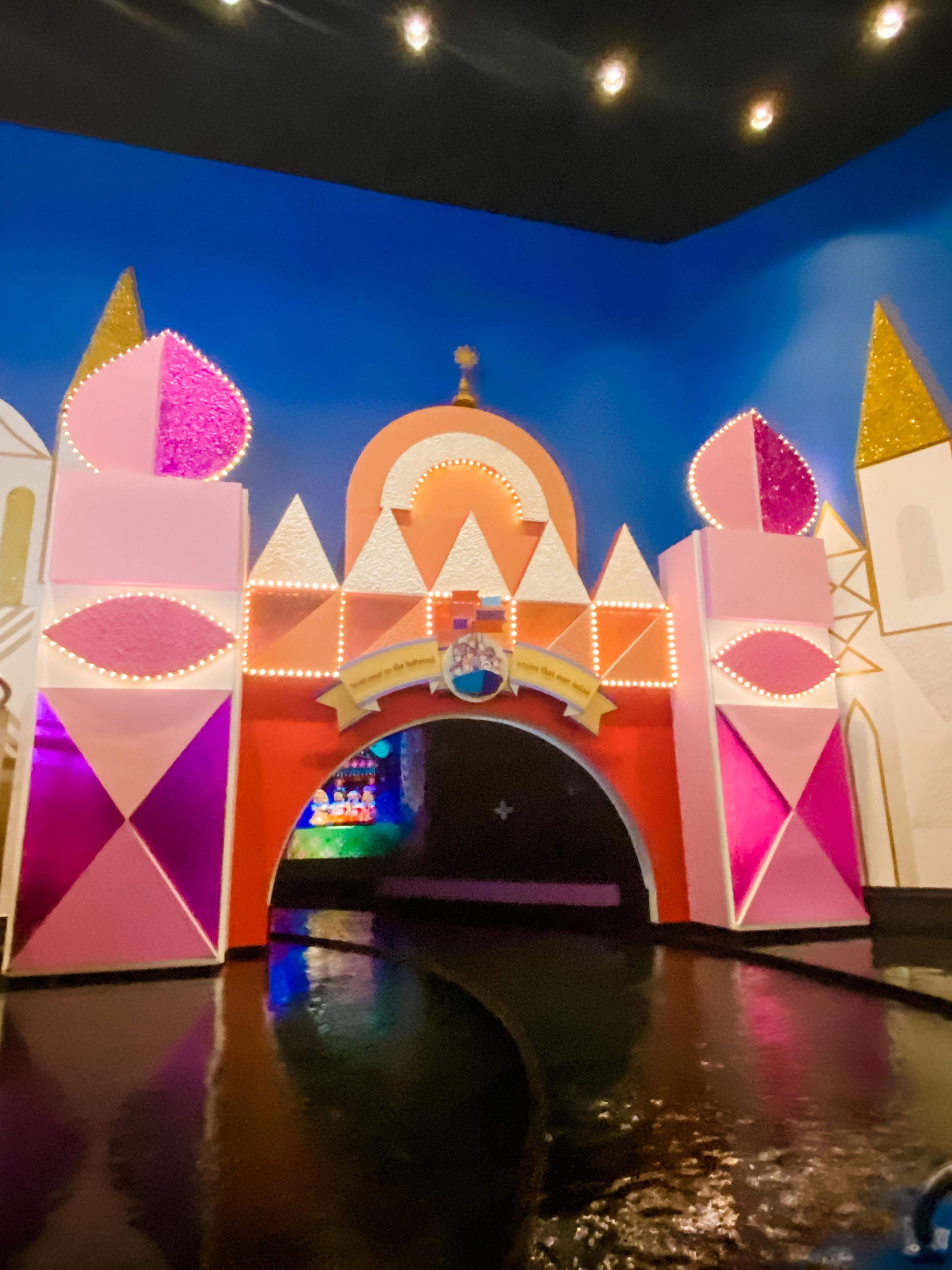 Colorful theme park attraction entrance featuring pink, orange and white castle-like architecture with geometric patterns and lighting.