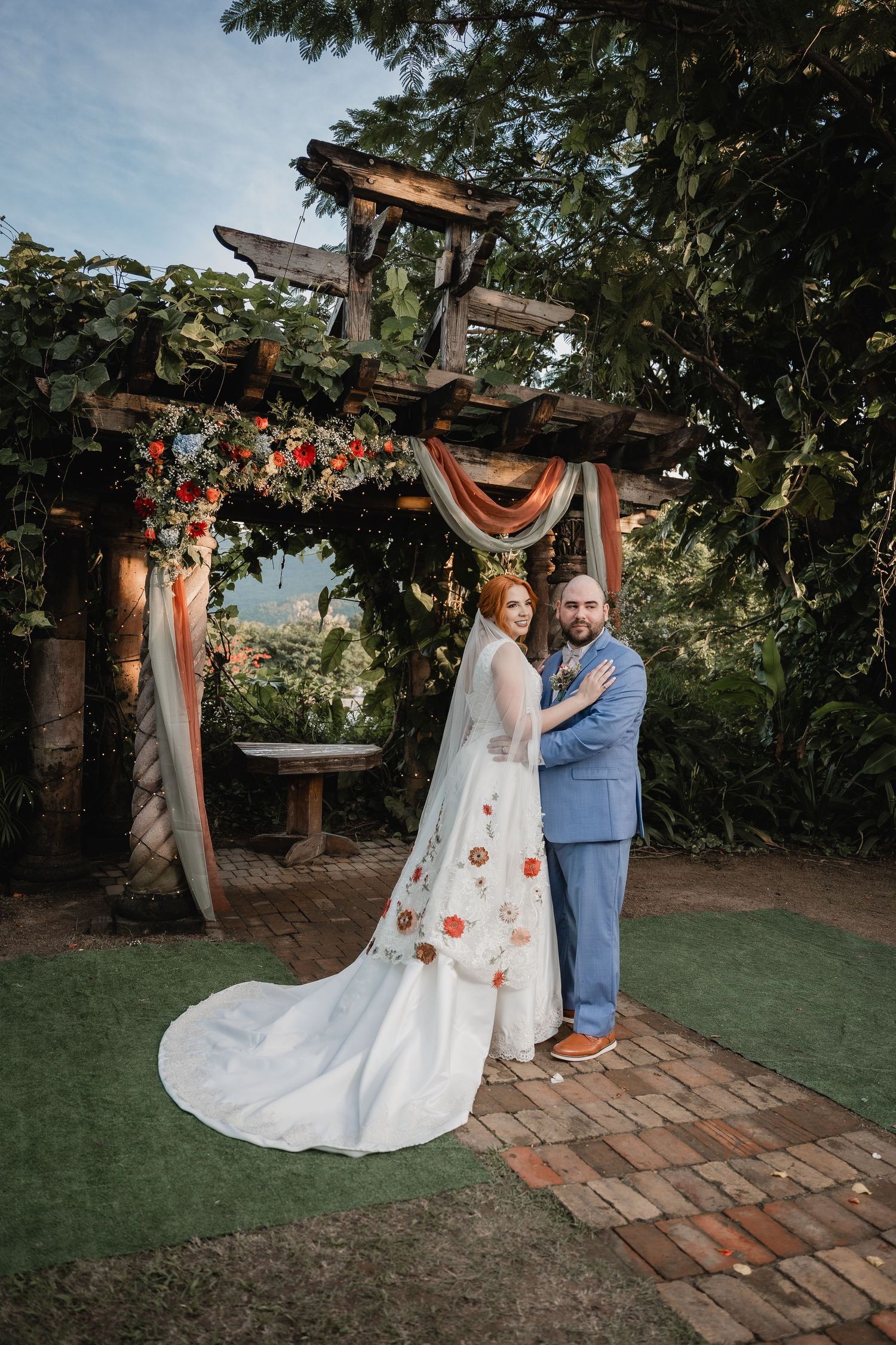 Couple posing under floral decorated wooden arch with orange and white draping.