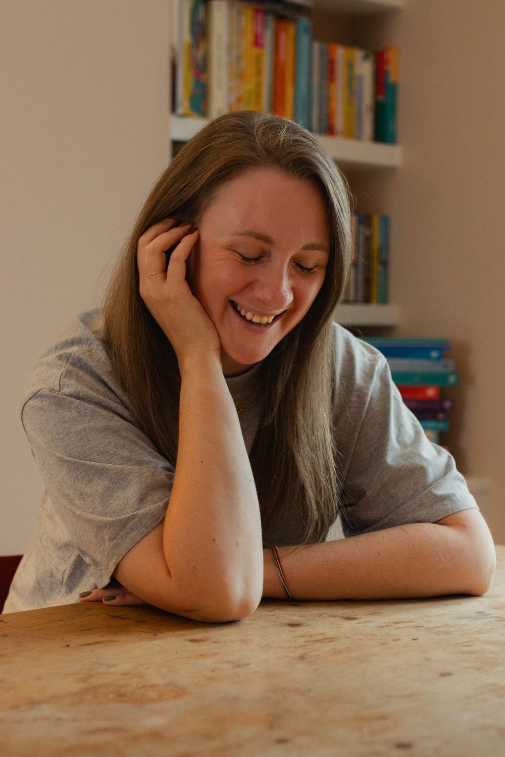 Person resting head on hand at wooden table with bookshelf in background.