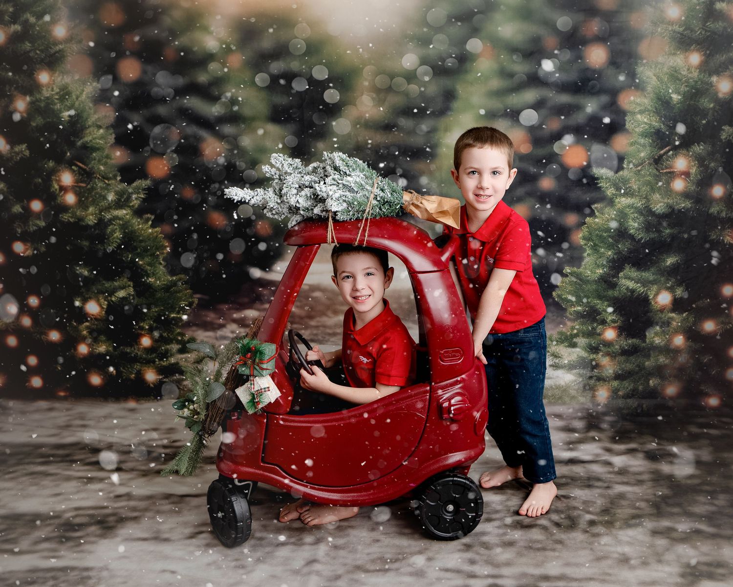 Two children in matching red outfits play with a decorated Christmas cozy coupe toy car amid falling snow effect.
