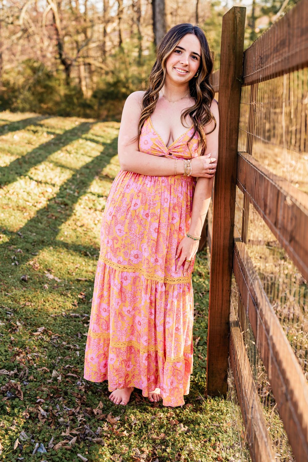 A pink floral maxi dress is worn on a High School Senior in a sunny outdoor setting with a wooden fence in the background.