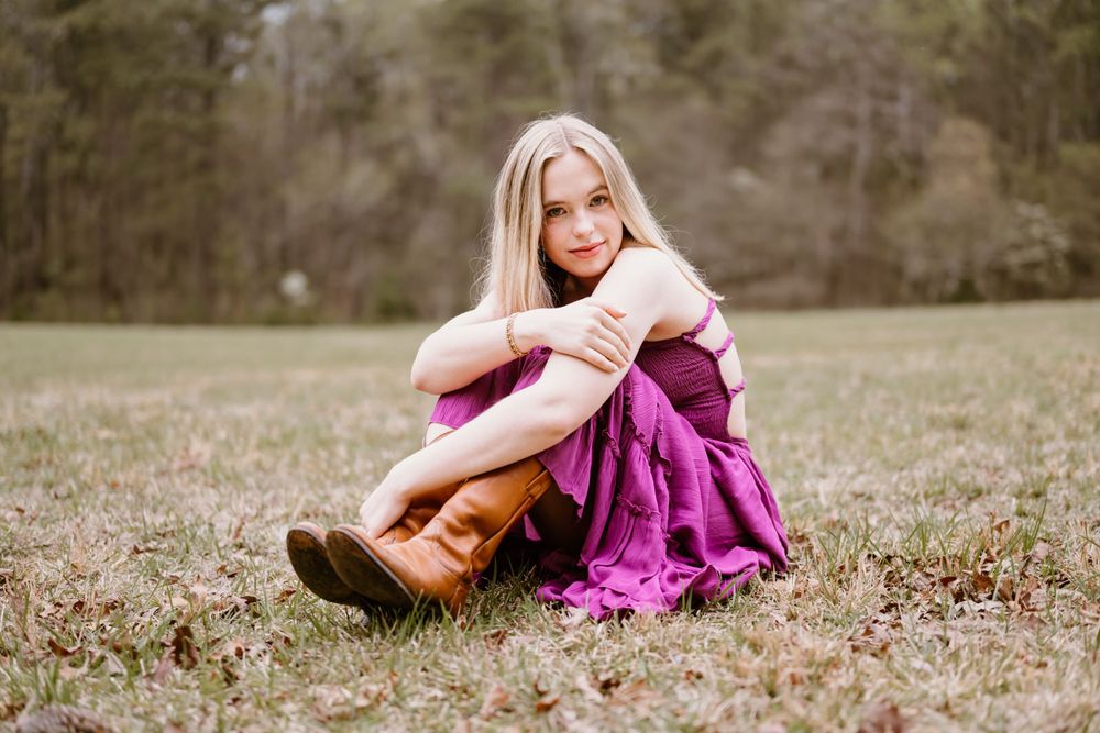 A High School Senior in a magenta dress sits on brown grass in a dreamy outdoor setting, wearing brown boots.