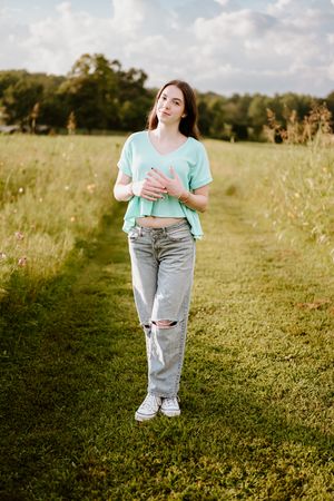In a sunny meadow, someone wearing a mint green shirt and ripped jeans walks through tall grass on a summer day.