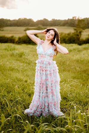 A figure dances joyfully in a ruffled pastel dress amid tall grass during a warm sunset in the countryside.