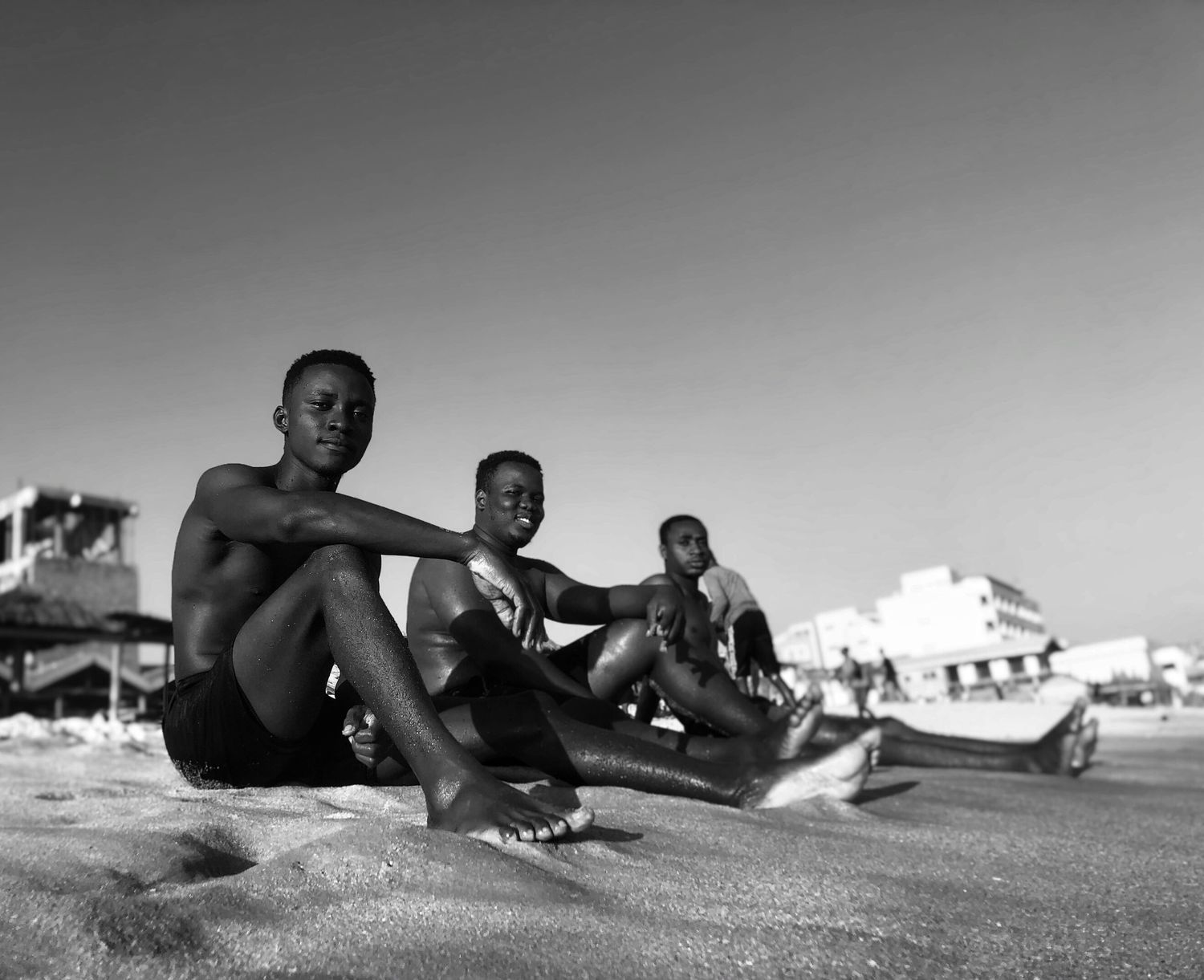 Three young men relaxing on a sandy beach under clear skies, captured by photographer Dennis Fofie.
