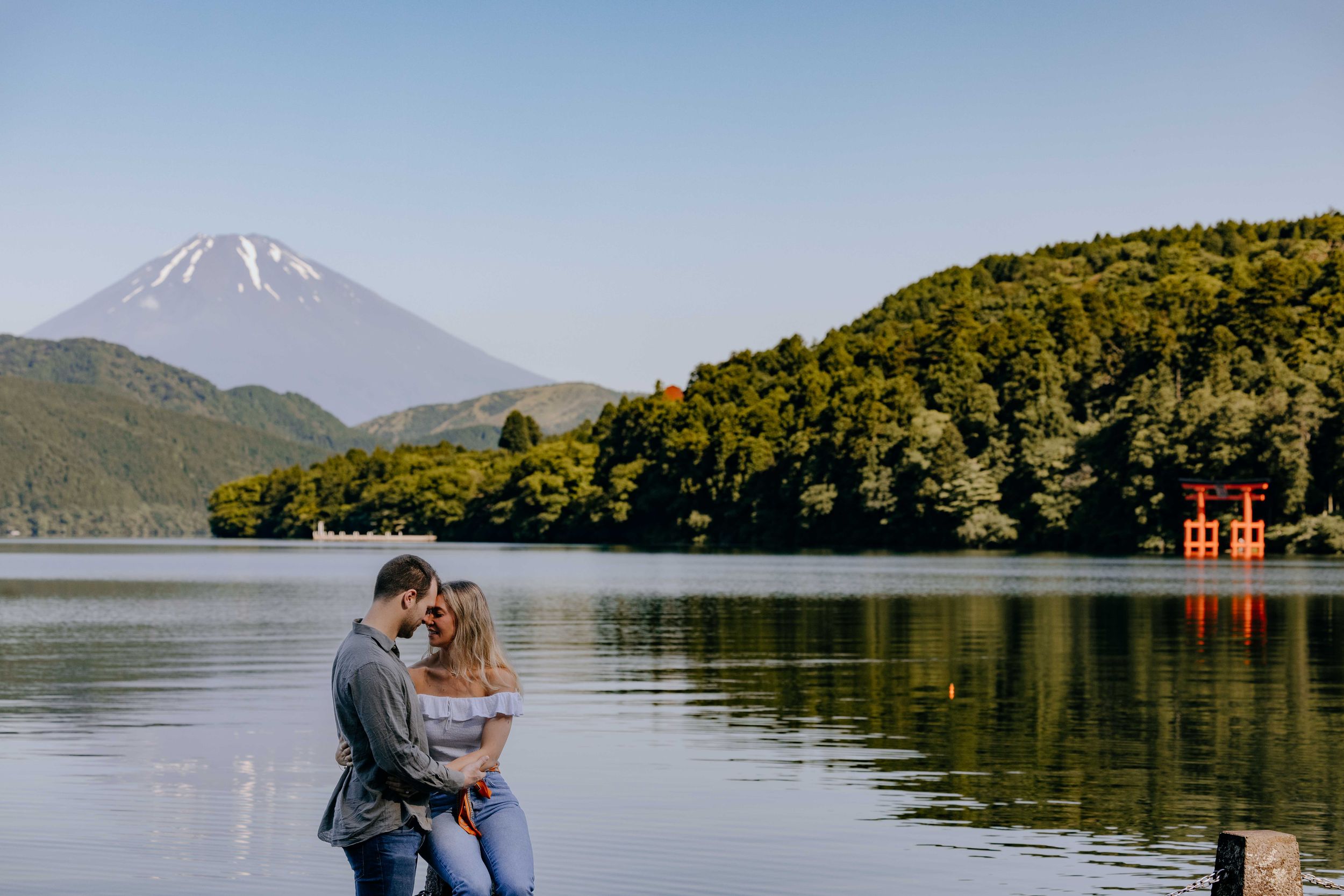 A couple sits by Lake Ashi with Mount Fuji and a red torii gate in the background in Hakone, Japan.