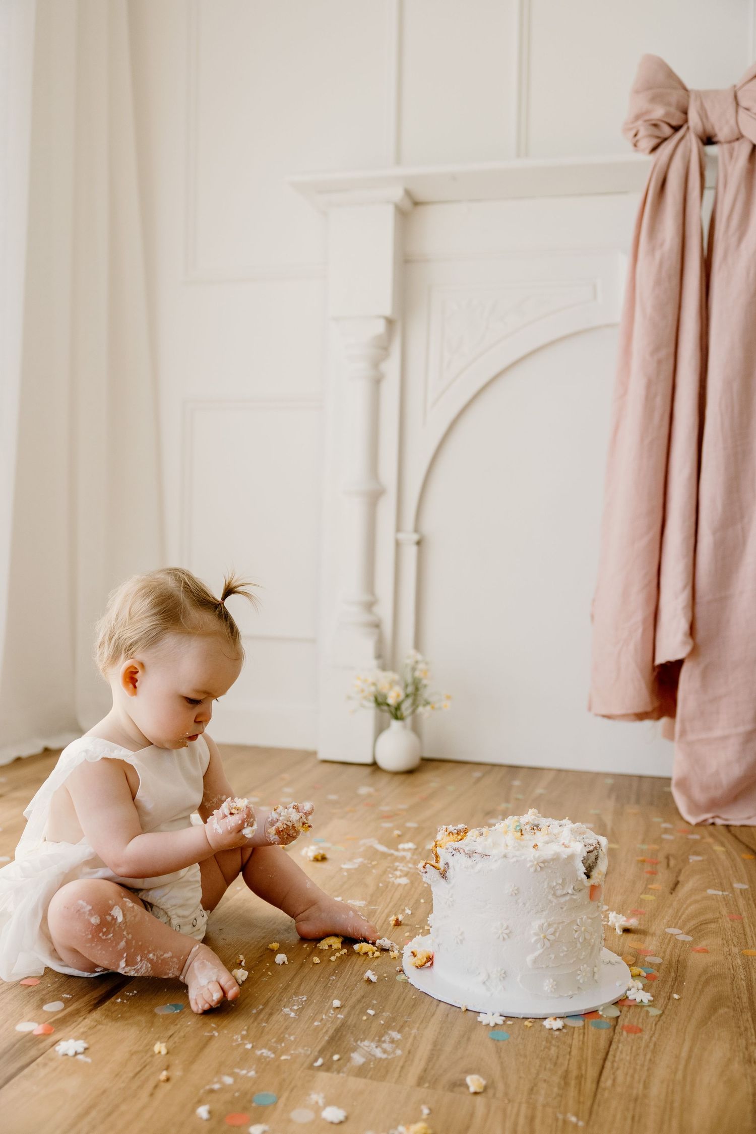 Small child in white outfit playing with cake smash setup on wooden floor.