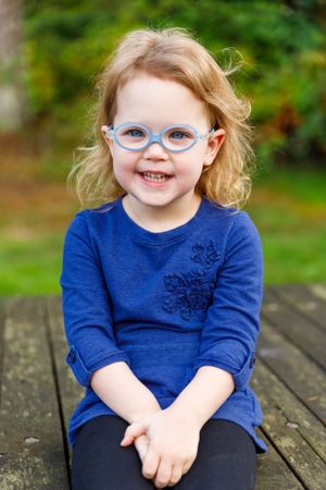 Portrait of a youngster in blue glasses and dress sitting on a wooden bench outdoors.