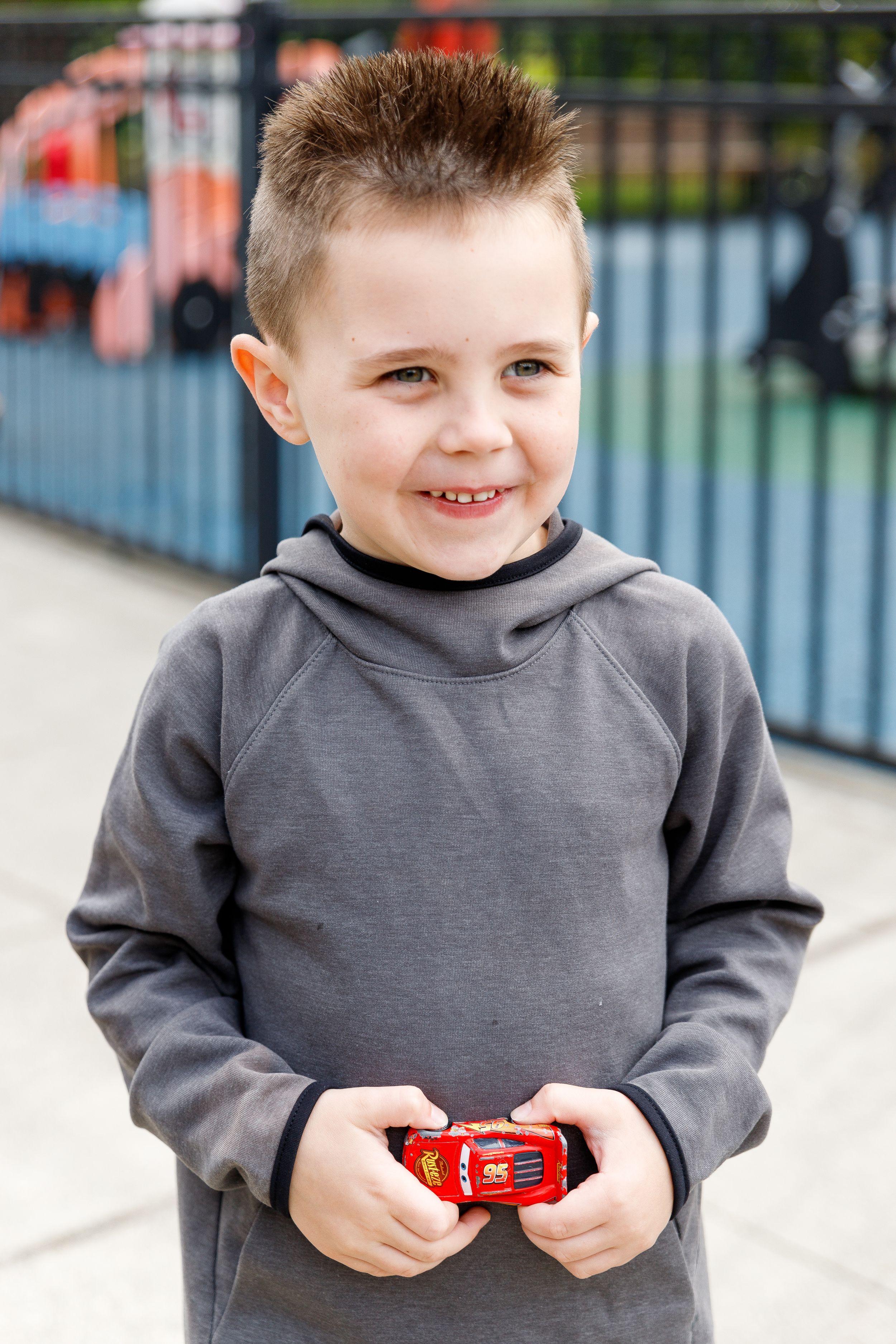 Young student in a gray hoodie holding a red toy car outdoors.