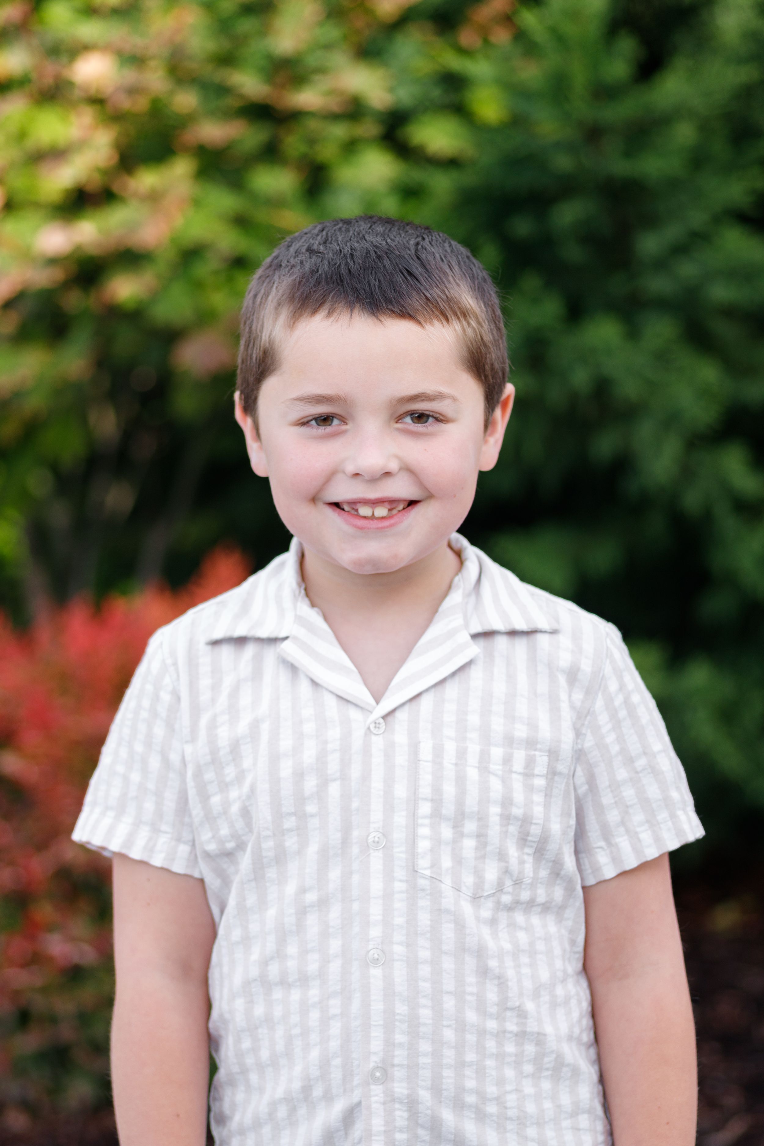 A young child wearing a white striped short-sleeve shirt smiles outdoors with autumn foliage in the background.