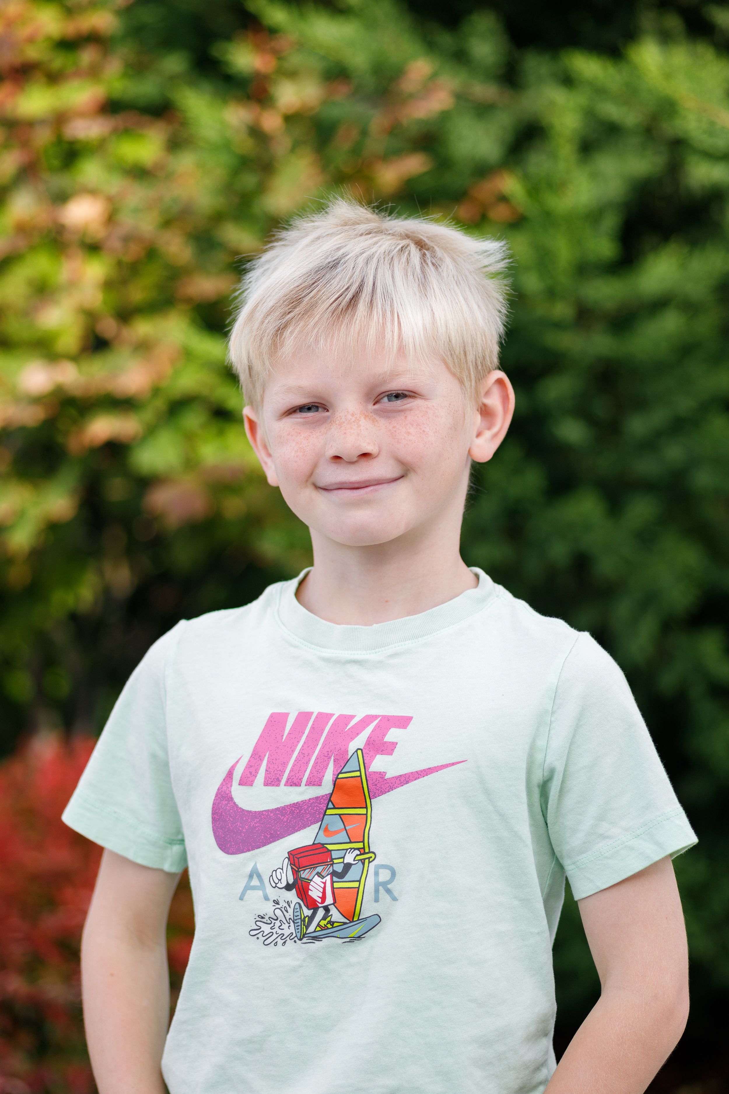 A child wearing a mint green Nike t-shirt with pink logo smiles at the camera in an outdoor setting.
