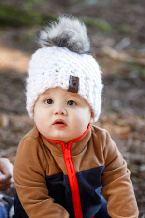 Baby wearing a white knit beanie with pom pom and brown colorblock jacket with red zipper.