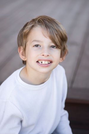 Portrait of a smiling child in a white sweater against a wooden deck background.