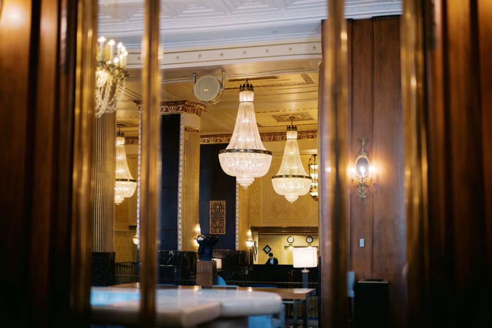 Elegant crystal chandeliers hang in an ornate room with dark wood columns and golden wall sconces.