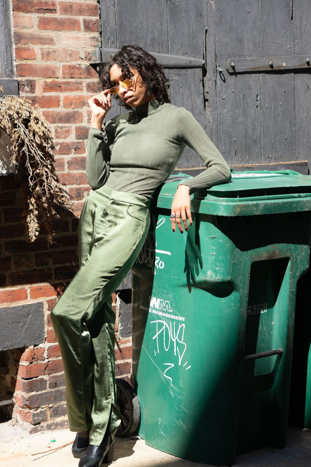 Street style portrait showing olive green monochrome outfit against brick wall and green dumpster.