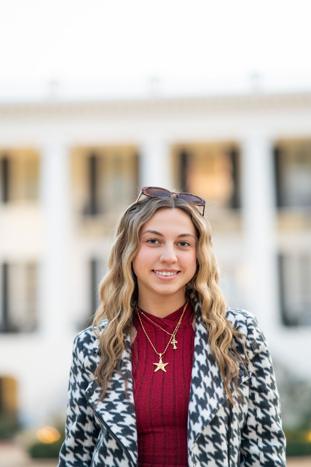 Young woman wearing a burgundy sweater and houndstooth pattern coat with sunglasses on head smiles outdoors.