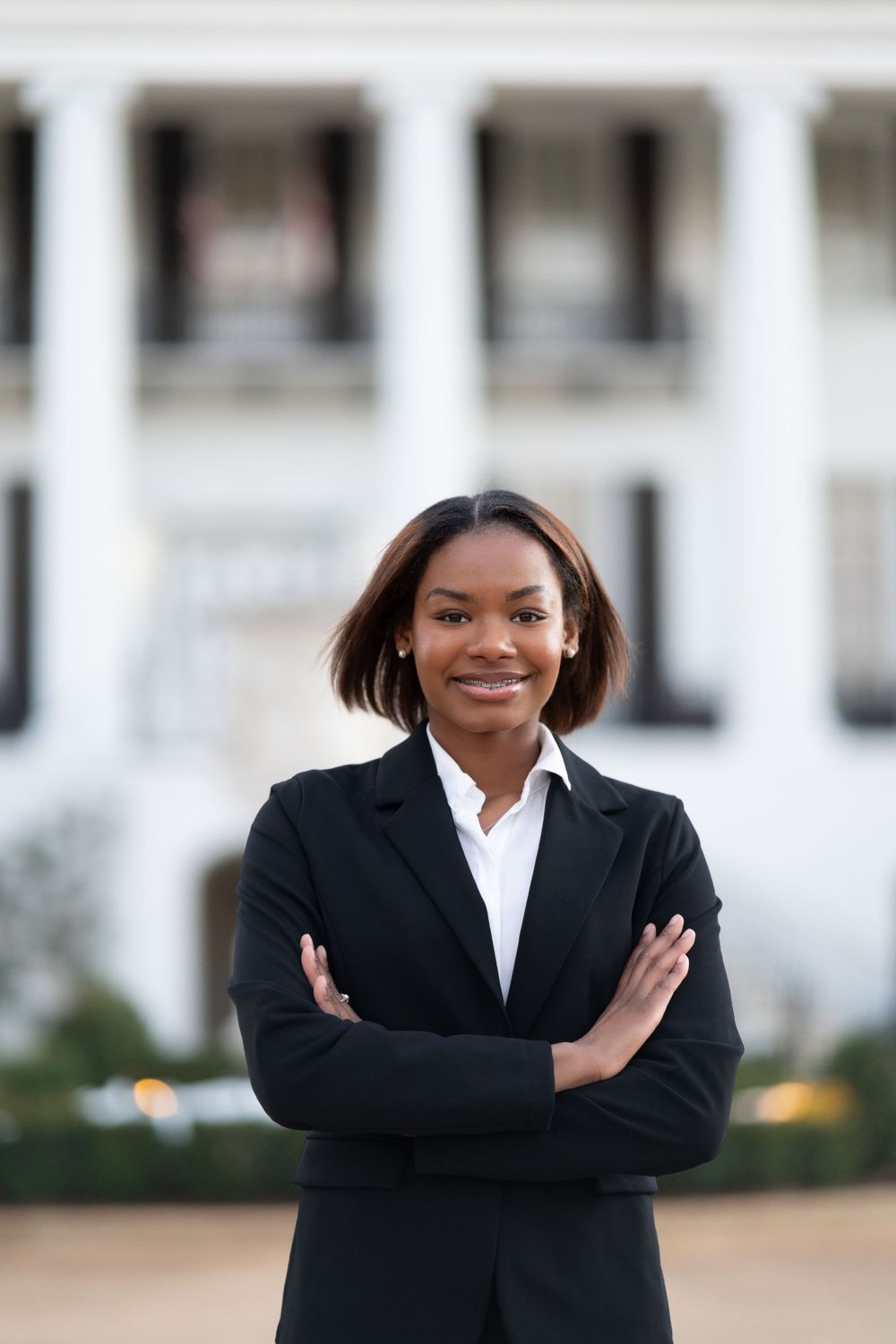 Professional in a black business suit and white shirt stands confidently with arms crossed in front of a columned building.