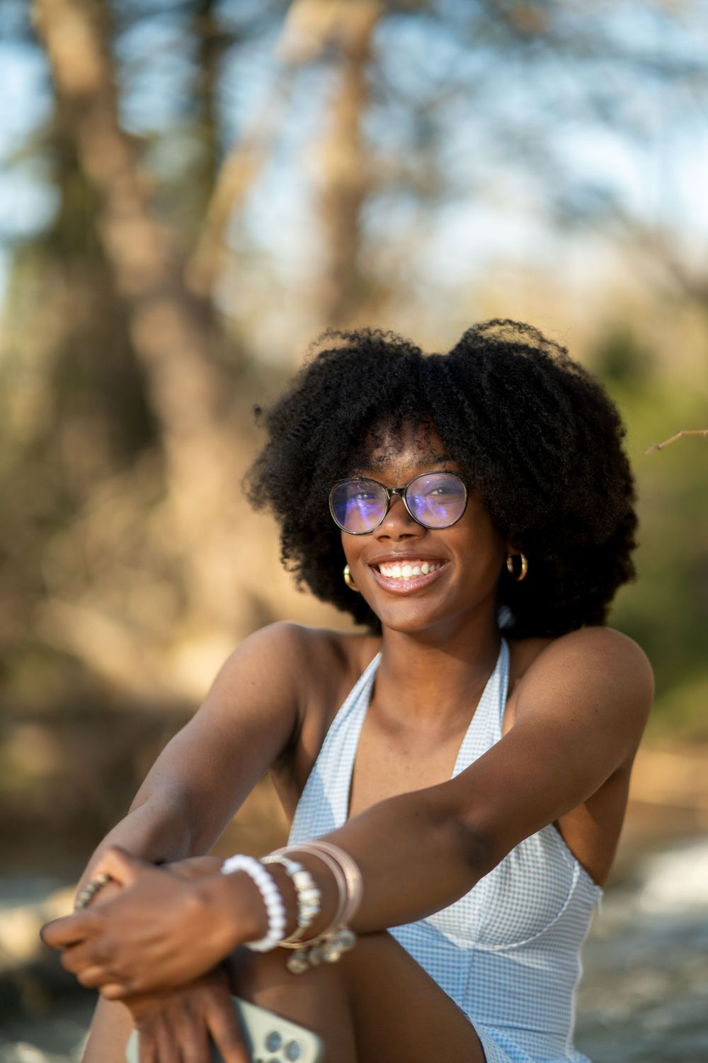 Person with natural curly hair wearing glasses and a white halter top poses outdoors in natural lighting.