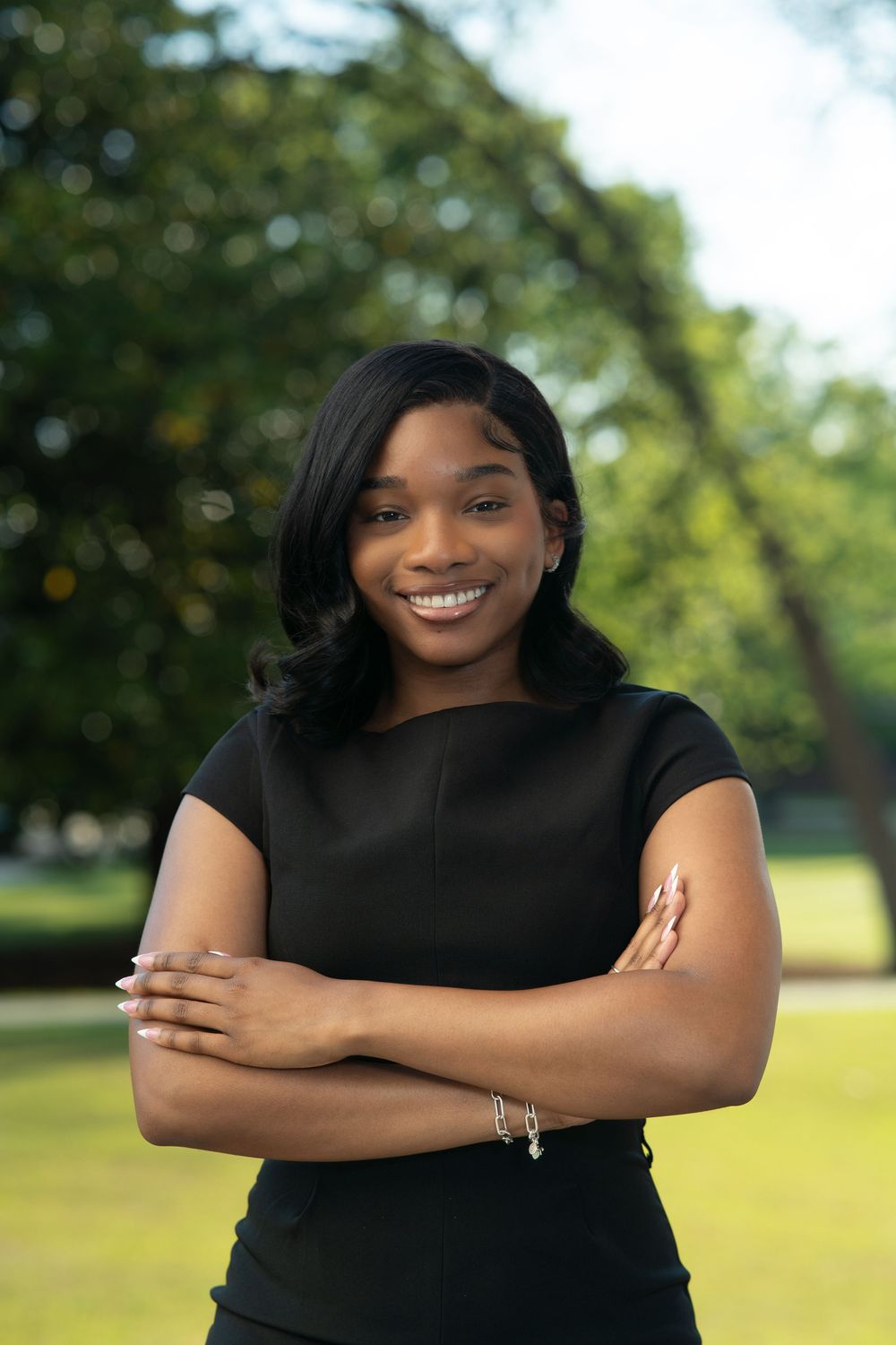 Professional outdoor portrait of someone in black attire smiling with arms crossed against a blurred green background.