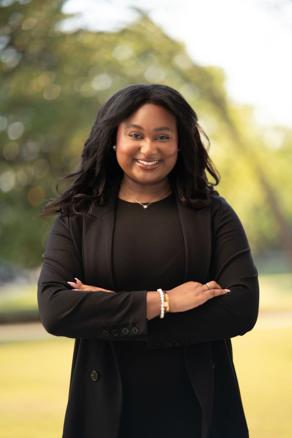 Professional portrait of someone in a black blazer and top standing with crossed arms outdoors on a sunny day.
