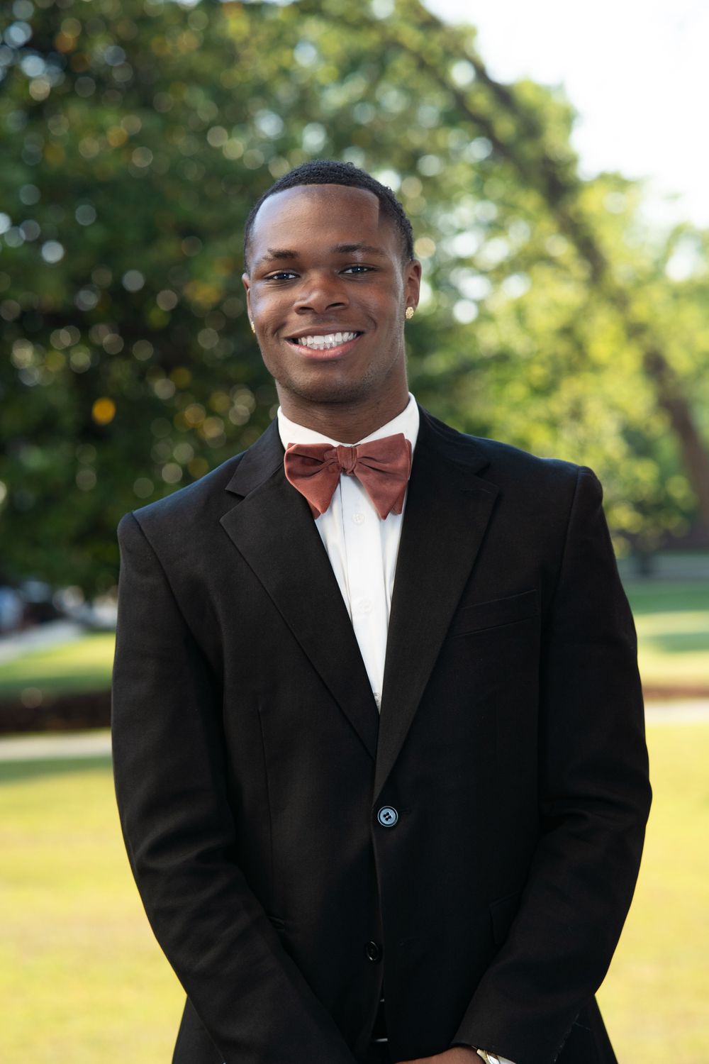A person wearing a black tuxedo and burgundy bow tie smiles warmly while standing outdoors in a garden setting.