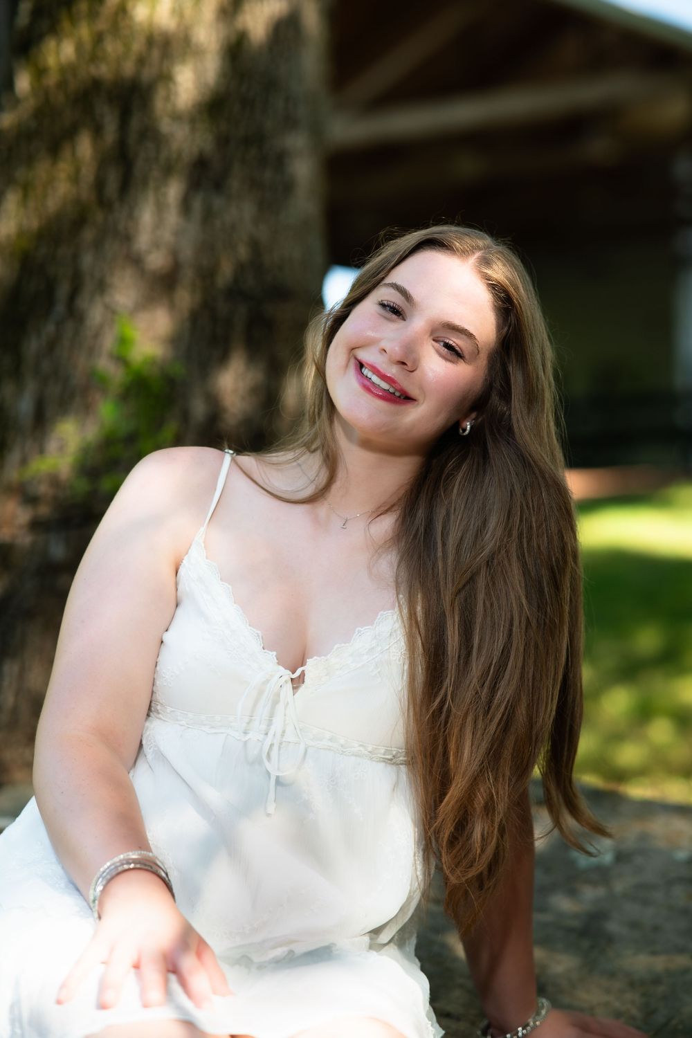 A joyful portrait in a white top poses outdoors with long brown hair and a bright smile on a sunny day.