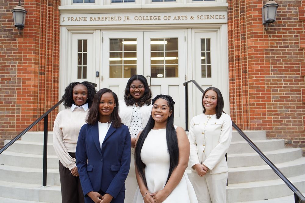 A group of students smile and pose together outside a brick college building with white columns and doors.