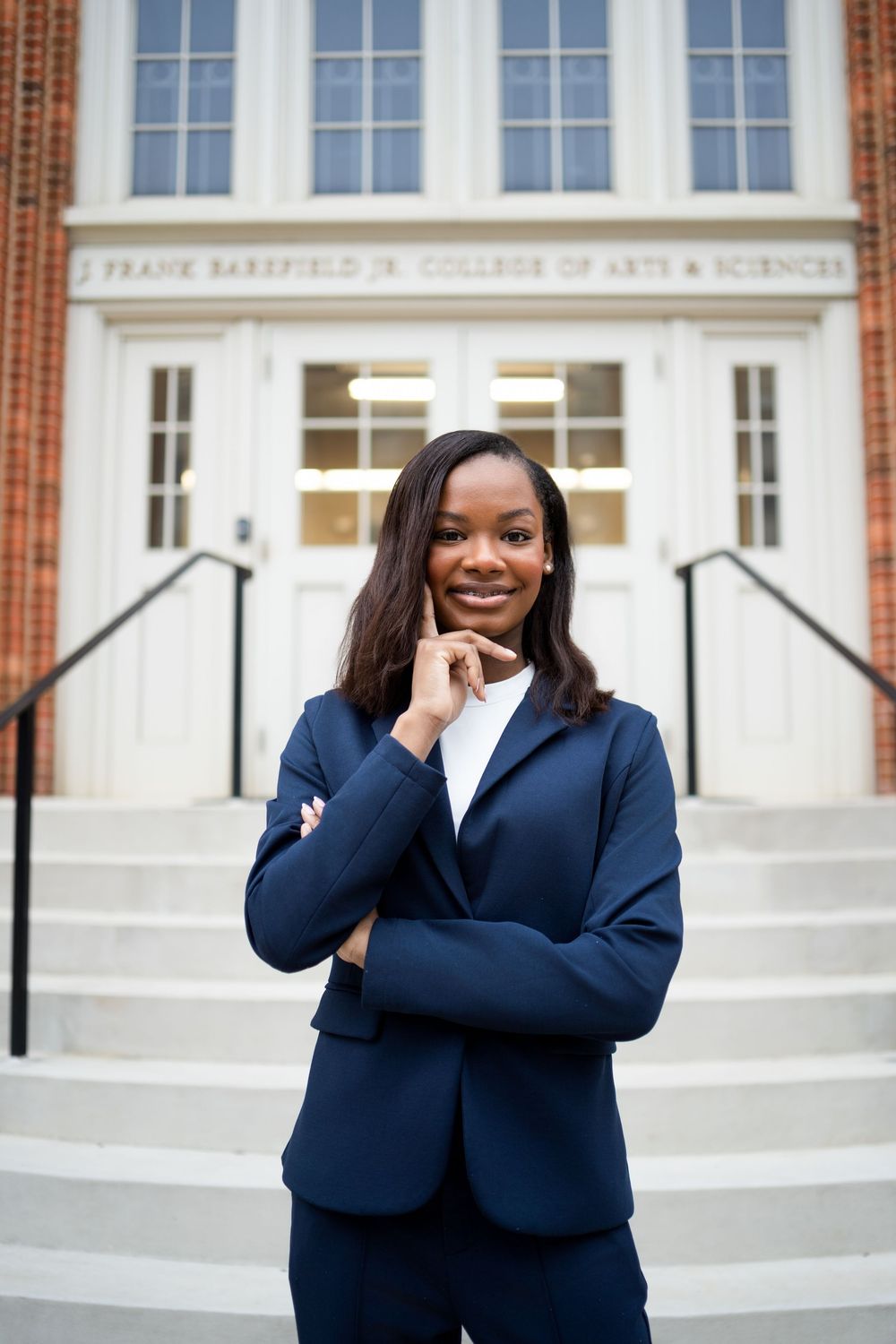 A college graduate in a navy blue blazer poses for graduation photos in front of a campus building.