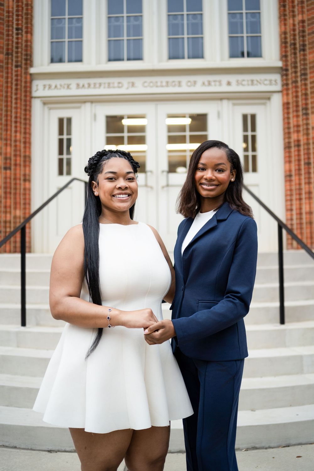 Two people in contrasting white and navy outfits pose together on steps outside a brick building.