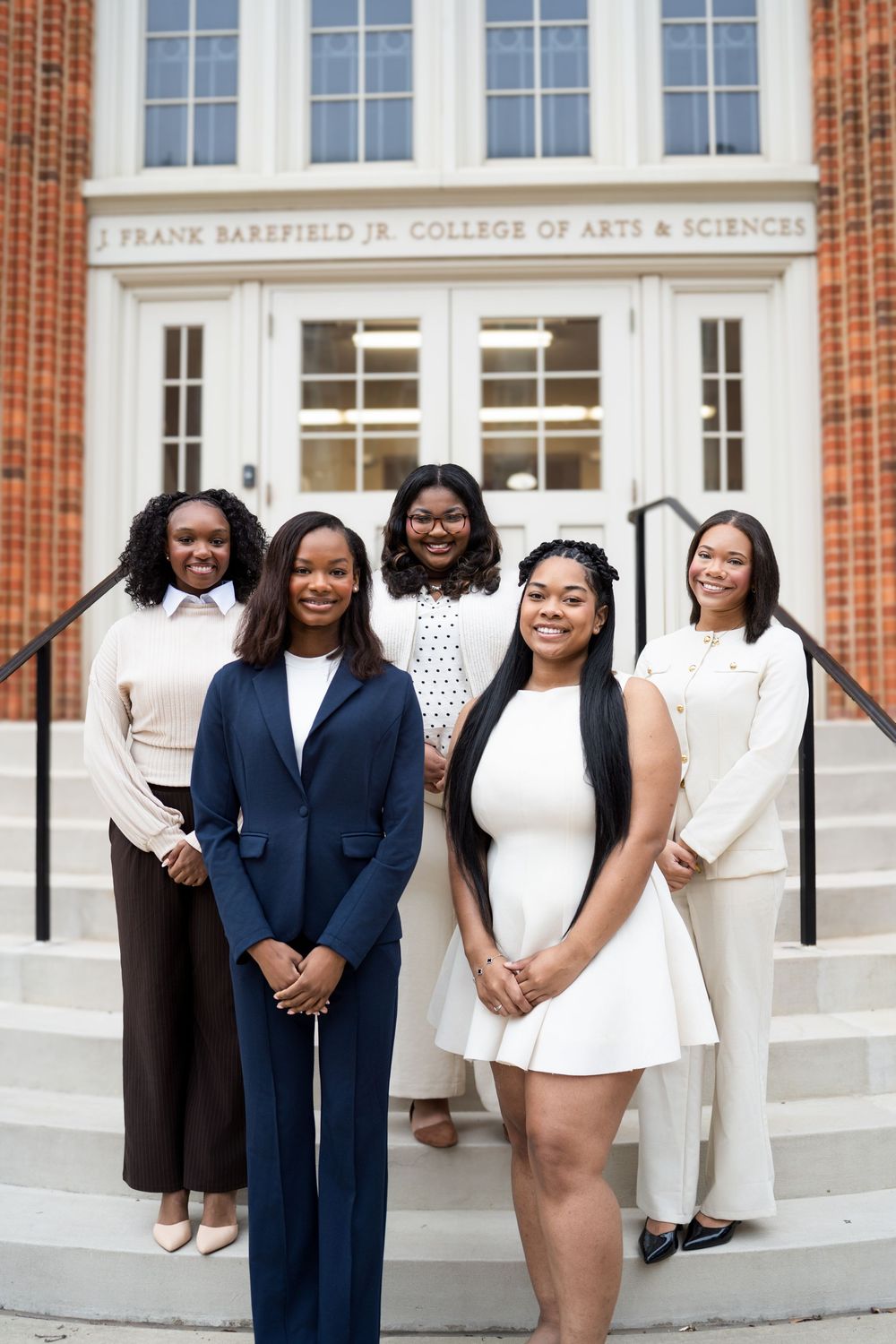 A group of graduates in navy blue and white attire pose together outside a college building.