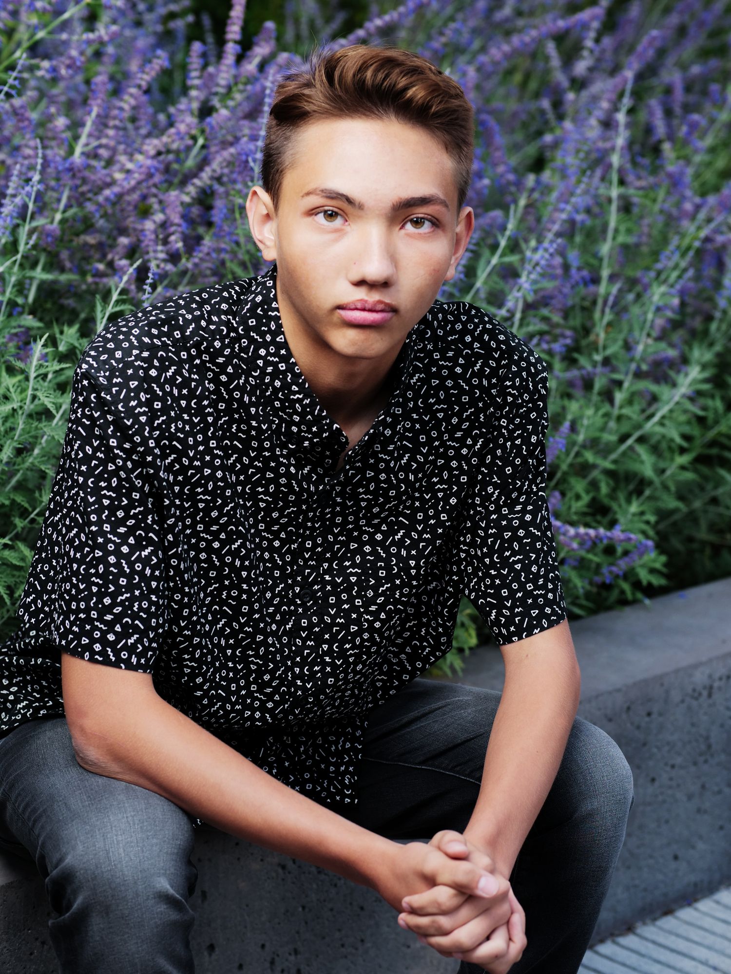 A senior in bold pattern shirt sits on a stone ledge surrounded by blooming lavender flowers in a garden setting.