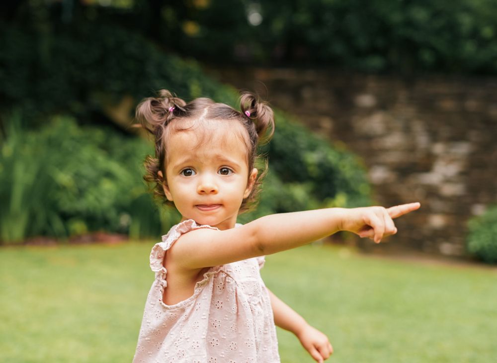 A small child in a pink dress points while standing in a lush garden setting.