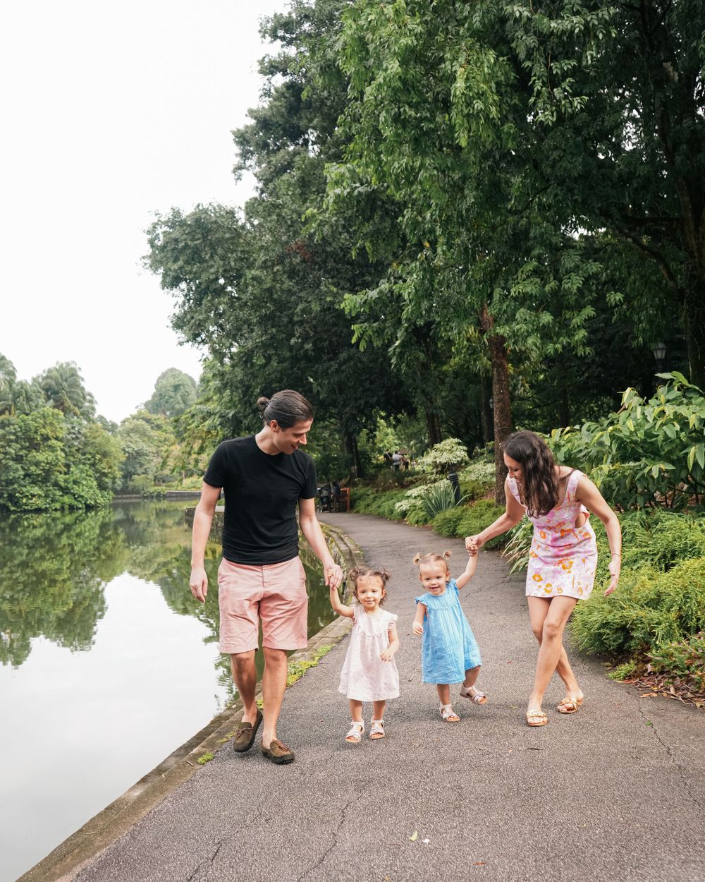 A family walks together along a peaceful pond path surrounded by green trees.