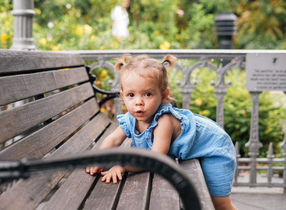 A toddler in denim explores wooden park bench with curiosity.