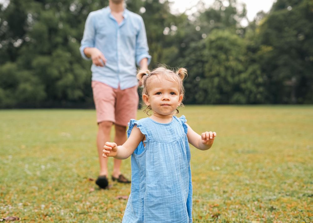 A toddler in blue dress toddles across grass while someone follows behind.