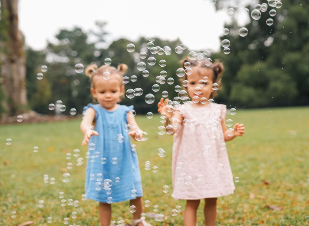 Two young children in sundresses play with bubbles on a grassy field.