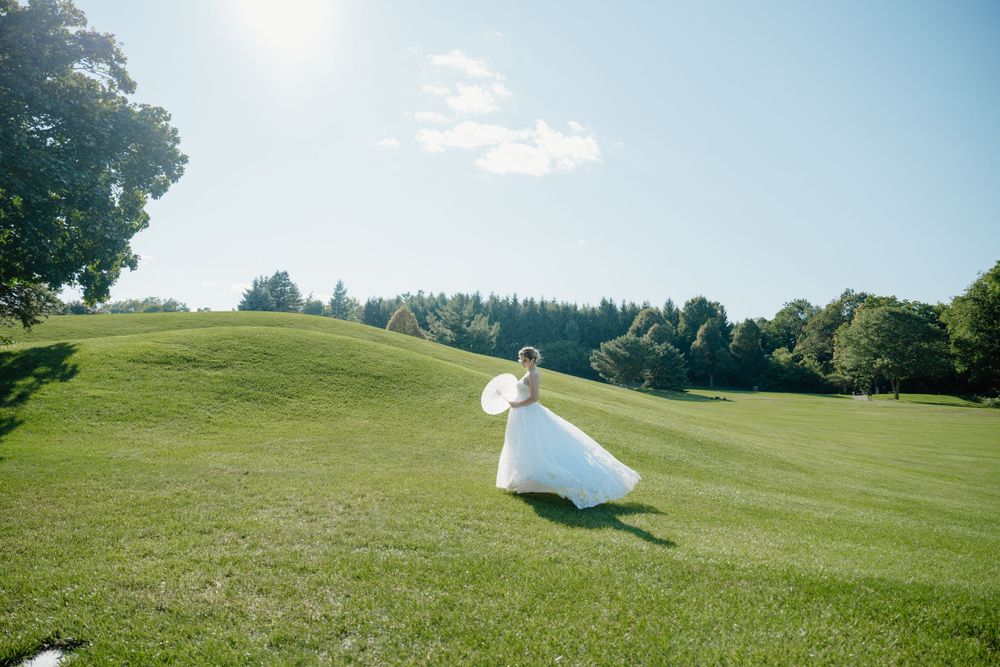 A figure holding a white umbrella stands on a lush green hilltop overlooking a scenic forest landscape on a sunny day.