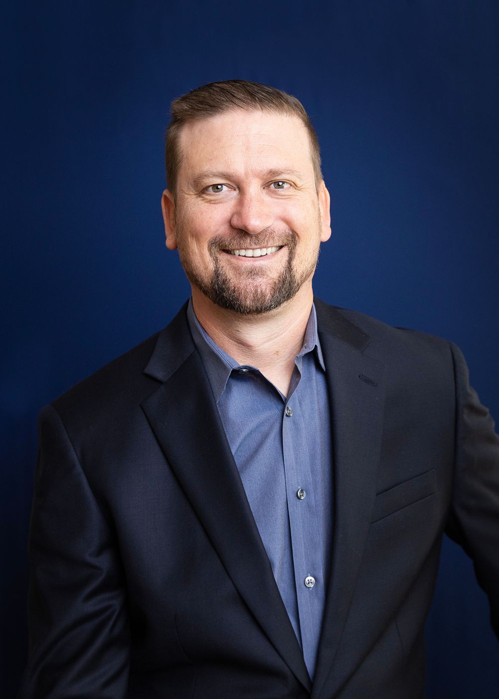 Professional headshot photograph showing a person in a dark suit jacket and blue collared shirt against a navy background.