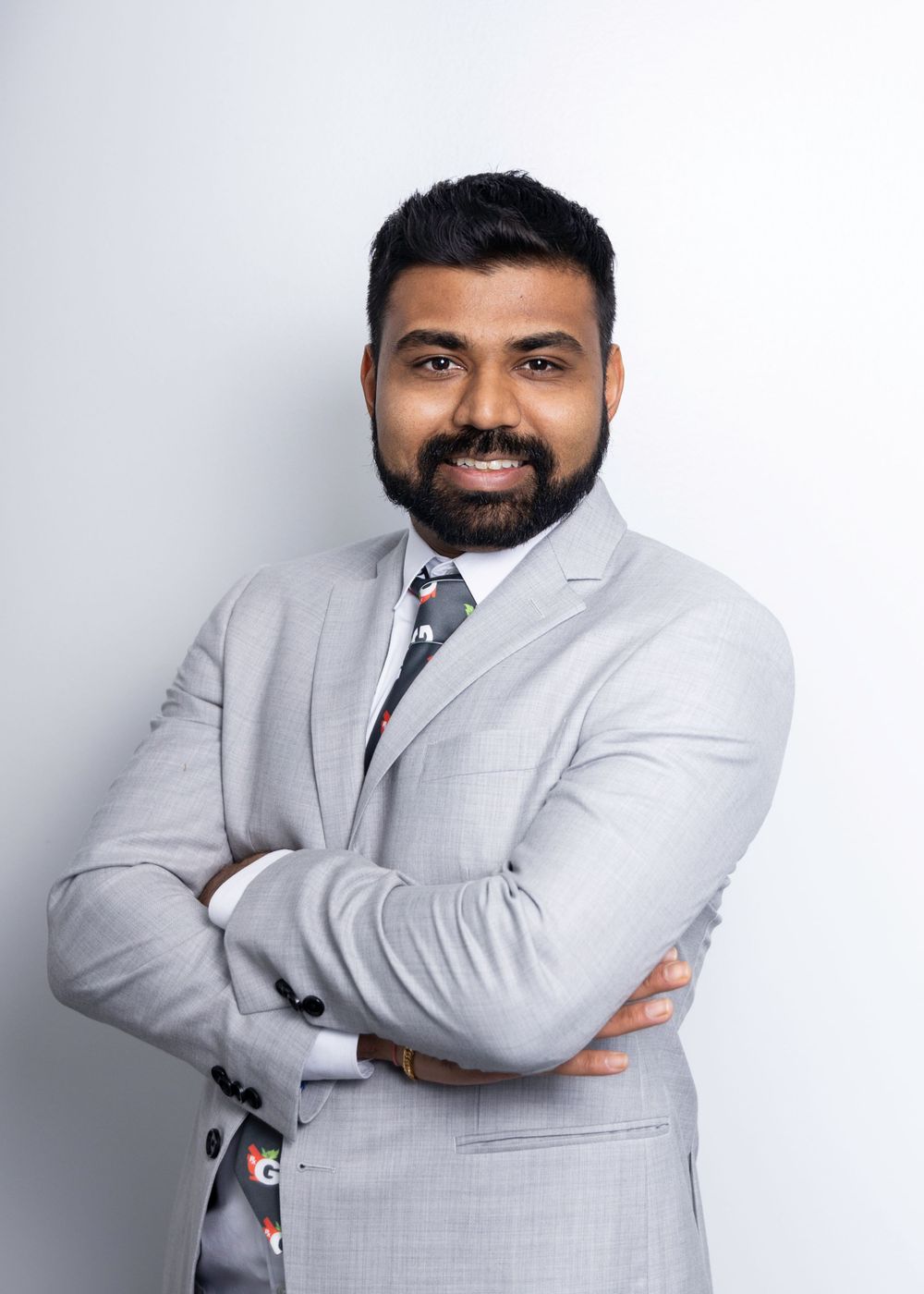 Professional portrait of someone wearing a light gray suit and patterned tie standing with crossed arms against a white background.